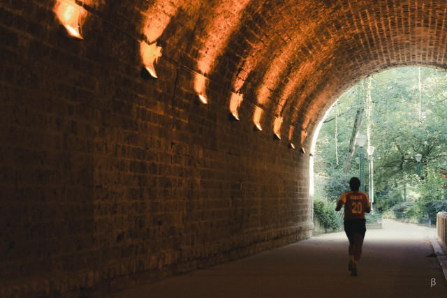 The image shows a person jogging through a dimly lit tunnel, likely made of brick or stone. The walls of the tunnel are textured and illuminated by warm lighting, casting a reddish-orange glow. The person is wearing an orange shirt as they move through the passageway, which appears to be part of a public space or park setting.