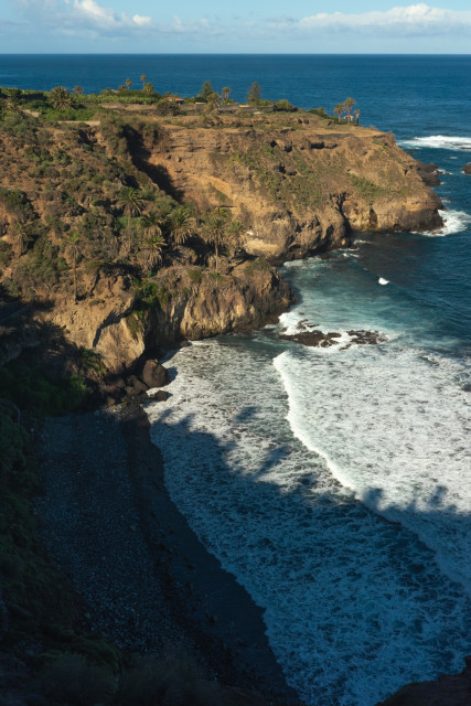 La vista de la playa de Castro desde el acantilado, con las paredes rocosas con palmeras y las olas golpeando fuerte.