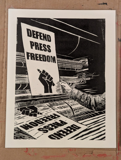 A black and white linocut print of a printing press in the process of printing a newspaper with the words “Defend press freedom”, and a fist holding a pen aloft. The arm of a pressman is visible, and they are holding up a print to inspect.