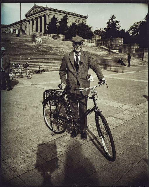 A dryntintype photo of a gentleman wearing tweed and posing in front of the Philadelphia art museum. 