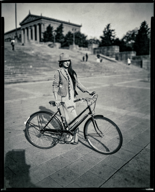 A black and white photo of a woman posing with a bicycle in front of the museum. A paper negative on 8x10. 