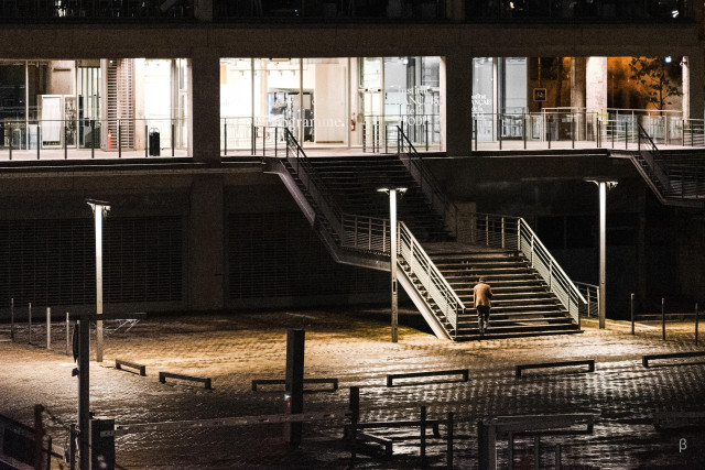 This appears to be an image of a public staircase leading up to a building or plaza at night. The stairs are surrounded by railings and illuminated by overhead lights, creating an atmospheric setting. A lone person can be seen climbing the steps, silhouetted against the lit building facade. The overall scene suggests a peaceful, urban environment.