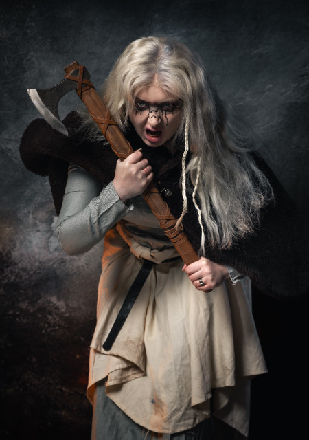 A person with white-blonde hair in a Viking outfit holding an axe stares and snarls at the camera, the background is like a grey stormy sky.