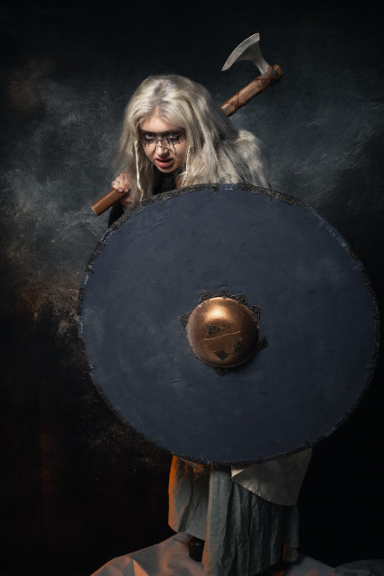 A person with white-blonde hair in a Viking outfit holding an axe and shield is looking over the shield at the camera, the background is like a grey stormy sky.