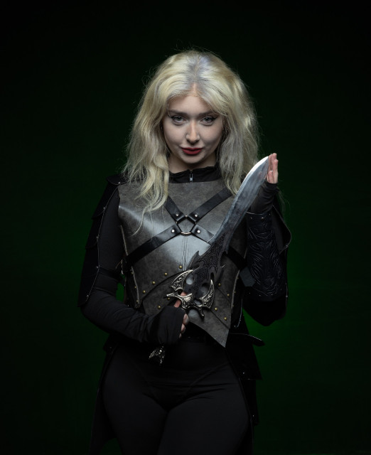 A person with white blonde hair in a black leather outfit holds a dagger and smirks. The background is dark and lit dimly with green light.