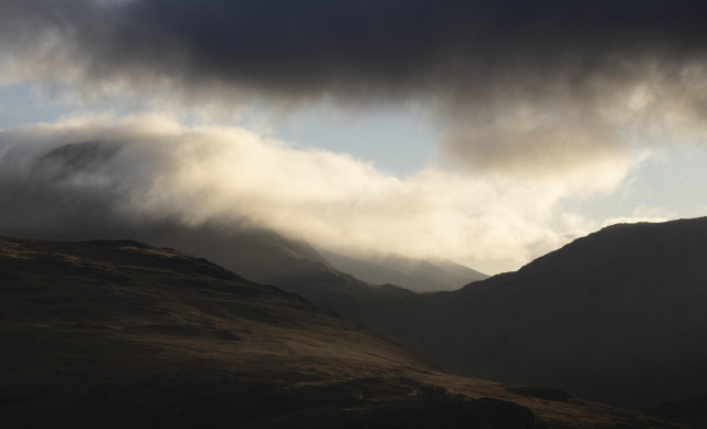 A mountain summit is partly covered by clouds. The clouds are lit up by late afternoon sunshine.