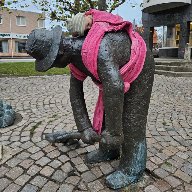 A bronze statue of a bent-over man wearing a hat is humorously wrapped in a pink baby sling, holding a doll with a striped cap. The playful addition of the baby sling contrasts with the laborious pose of the statue, symbolizing the dual demands of work and caregiving. This intervention is part of Arena Idé's International Men’s Day campaign, highlighting the unequal distribution of parenting responsibilities in Sweden. The scene is set in a cobblestone plaza with nearby buildings and trees, emphasizing the campaign's goal of sparking public reflection on modern fatherhood and shared caregiving roles.