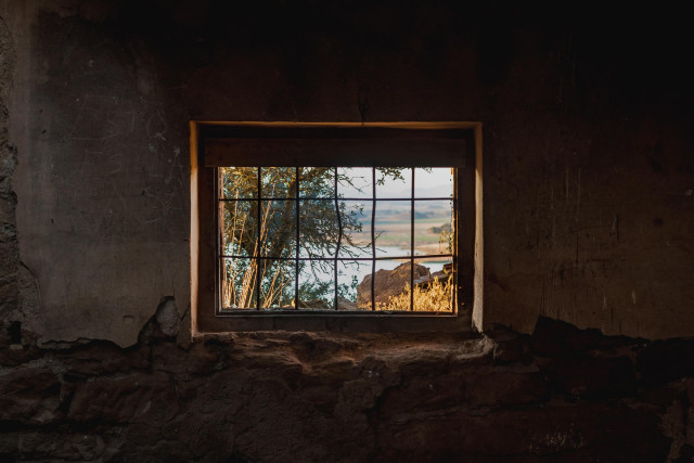 Se puede ver la pared oscura y raída en la parte inferior de un edificio abandonado. En el centro, una ventana con unos finos barrotes de rejilla, a través de la cual se ve parte un entorno natural.