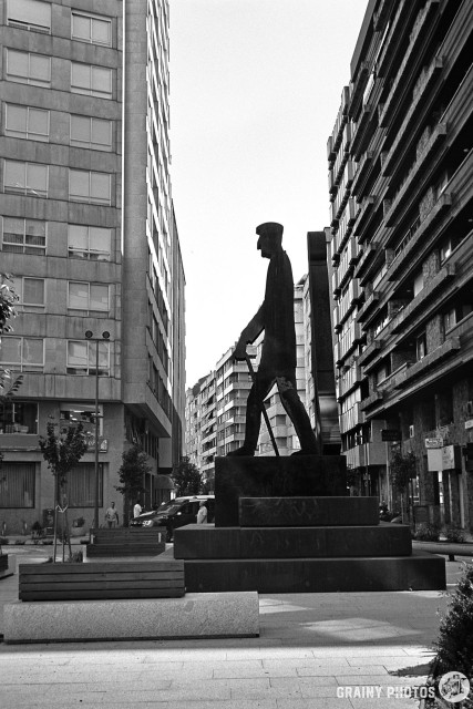 A black-and-white film photo of a steel sculpture in a pedestrianised area of a man walking. The figure is silhouetted against the sky and blocks of flats.