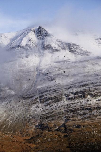 A colour photo of the mighty Liathach Mountain in late morning sunlight with some mist clinging to the upper reaches and red / brown vegetation at the bottom. A dusting of snow reveals the dramatic combination of rocky outcrops, scree slopes, debris slides and erosion channels. In the foreground, four crows fly across the scene. Torridon, WesterRoss, Scottish Highlands.

Photo by and copyright of Paul Henni.