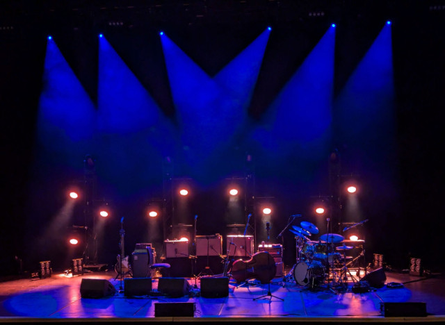 Photo of a stage illuminated by six deep blue spotlights shining from above and eight less powerful but warmer lights from the back. The blue and amber light reflects and casts purple-pink shadows across the stage from a bank of amps, a drumkit to the right, a double bass to its left and a guitar on the opposite end. There's a row of stand microphones and more boxy equipment across the middle.