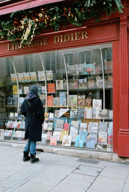 A person in a long black coat, wooly hat, and boots standing on a pavement outside a big glass bookstore window. The window is framed in a red wooden front with gold lettering on top reading 'Librairie Didier', and above the sign, there's a stretch of green pine branches and red and gold decorations.