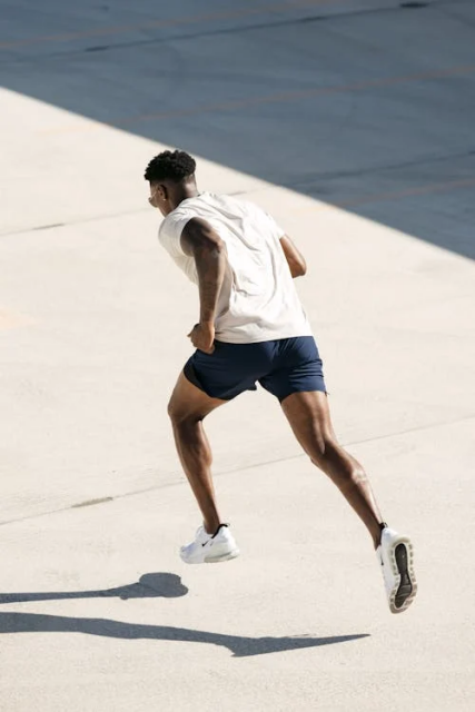 A man in white t-shirt, navy shorts, and white trainers, mid-sprint across a concrete surface. His muscles are tense and it's a very athletic pose.