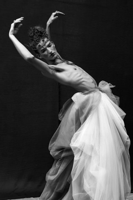 A soft black and white photo of a person with a bare chest and flowing white net ruffles from the waist down, stretching backwards with arms up, looking down to the side. They're wearing heavy ornamental jewelry around their forehead and neck. 