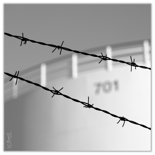 A black and white square image of the top of a fuel depot behind two rows of barbed wire which go diagonally throu the picture. The out of focus tank has the number 701 on it and there is a railing at the top. The sky is clear.