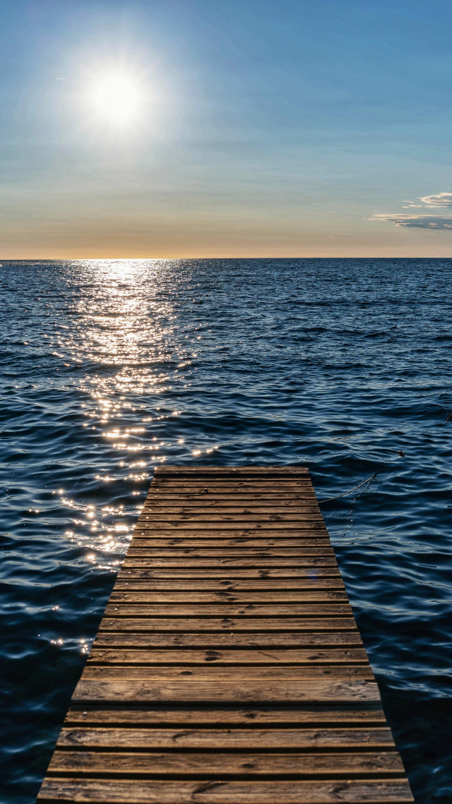 Ein Steg ins Meer hinein. Man sieht bis zum Horizont das Meer. Darüber die Sonne am fast wolkenlosen Himmel. 

A jetty into the sea. You can see the sea as far as the horizon. Above it, the sun in an almost cloudless sky.