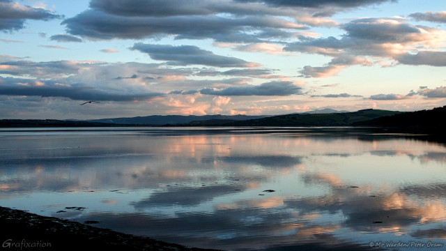 A photo of a wide lake under a cyan sky, with scattered and puffy clouds which the sunset is tinting to shades of peach and pink. On the horizon is a line of dark mountains, the foreground is the lake's gravelly edge. The slightly ruffled water is reflecting the sky, and a bird is flying away at eye level. It's calm and serene.