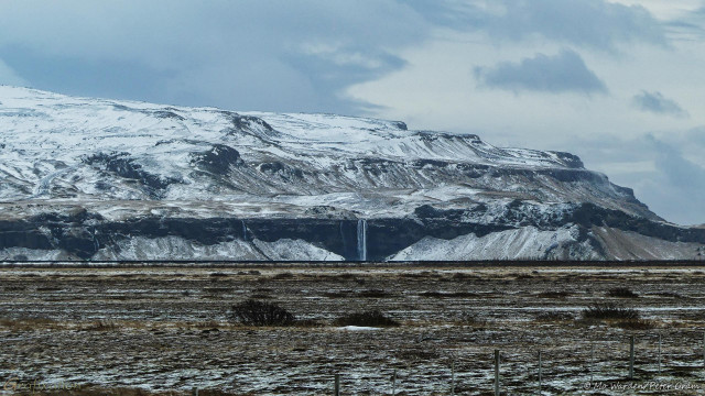 A winter photo of a mountainside of dark rock covered in ice. The foreground is withered brown vegetation dotted with snow. Near the base of the cliffside, the rockface abruptly flattens into a vertical face, it's foot debris-strewn. Three waterfalls can be seen; on the left is the top of a cascade but the base is hidden, left of centre is a small cascade, and near the centre is an impressive horsetail falls.