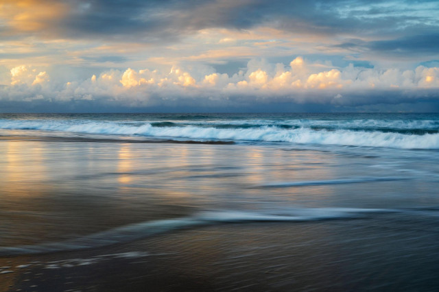 sunset seascape with some colourful clouds in the back reflecting in the wet sand. Foreground shows some moving water making lines and building the composition.