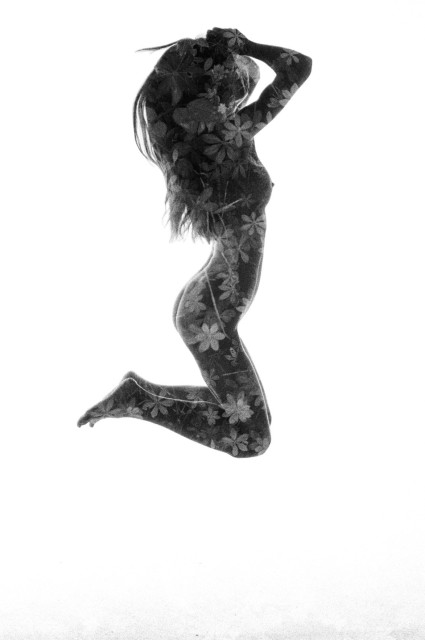 A naked woman is jumping in the air, legs bent. Her body is covered in flowery leaf texture from the double exposure. Black and white. 