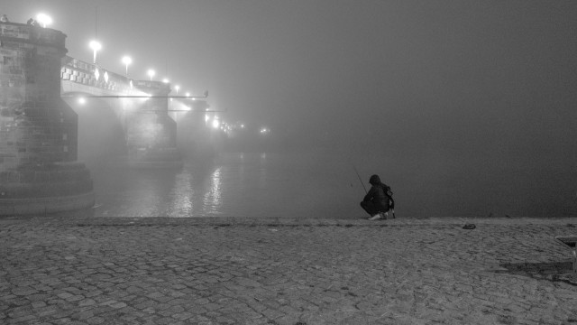 Nächtliche Schwarz-weiß-fotografie von einem gepflasterten Ufer vor einem Fluss. An der Kante des Ufers kauert ein Mensch in einer schwarzen Jacke mit einer Angel. Dahinter ist ein Fluss zu erkennen, über den eine Brücke verläuft. Das andere Ufer verliert sich in Nebel. Auf der Brücke sind Straßenlaternen, die die Szene in ein weiches, helles Licht tauchen.