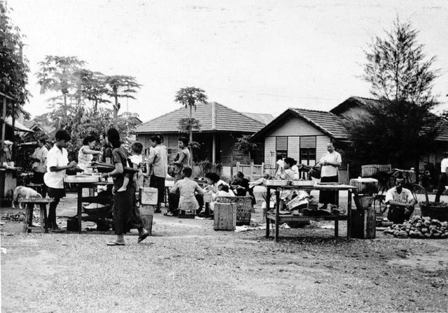 An outdoor market scene in rural Malaysia featuring several people engaged in various activities related to food preparation and sales. Children are present, some assisting adults, and others playing. The background shows simple residential houses and trees, conveying a communal atmosphere.
Black and white photo.