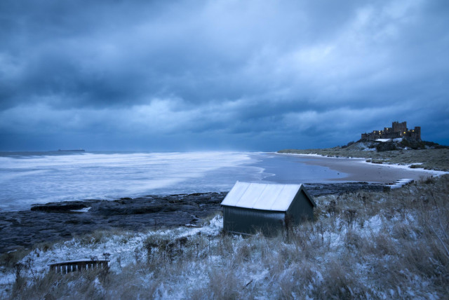 A photo taken in the blue hour, just before sunrise. In the foreground a beach hut and bench are covered in snow. Beyond this rocks give way to breaking waves and the beach. To the right of the beach a castle stands on a hill with lights visible in the early morning gloom. on the horizon an island is visible with a single light from the Farne (island) Lighthouse. 