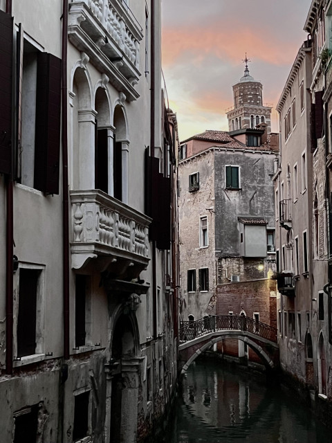 Colour photo taken looking down a narrow curved Venetian canal, with four-storey grey-white stone buildings on either side, a little bridge at the end before the canal turns a corner, and the crown of a slightly listing church bell tower peeking over the far rooftops. The water in the canal is a dark oily mossy green colour, and the curve of the bridge is clearly reflected in it. The light in the picture is quite unusual - there is no direct light on the buildings and they all look quite sombre and pallid, but the sky above is absolutely luminous, with two bands of white-edged pink clouds against pale blue, like a glowing Trans rights flag over the city. 