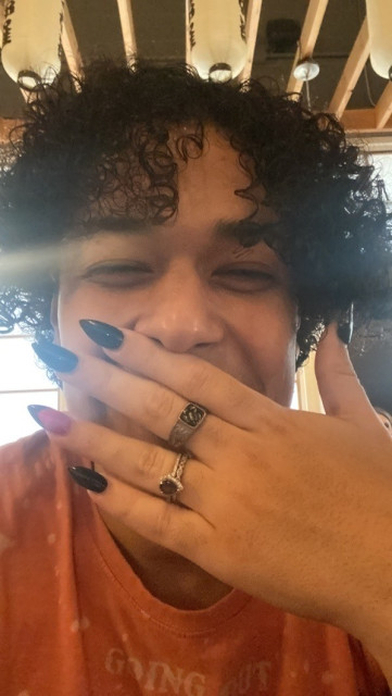 My comrade, Summer, stifling a laugh behind one hand. She has curly black hair & her fingernails are painted black & purple. She's wearing several rings & an orange t-shirt.