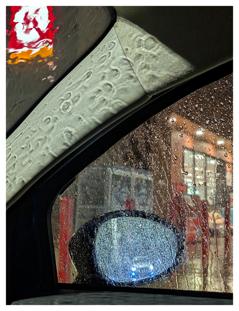 before dawn, in the rain. view of a storefront from the driver's seat of my fiat. through the windshield and passenger window a vehicle with blue fog lights at a fuel pump is seen on in rearview mirrow.