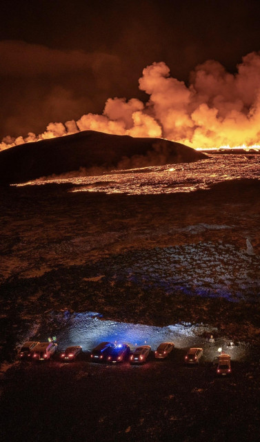 A line of cars with their lights on pointed toward a big lava field with a mountain and lots of smoke. A couple of people are standing close to the cars.