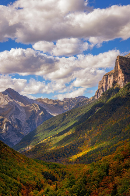 Fotografía vertical donde se ven un par de formaciones montañosas con faldas de vegetación con colores de otoño. En la parte superior se pueden ver un cielo azul con nubes.