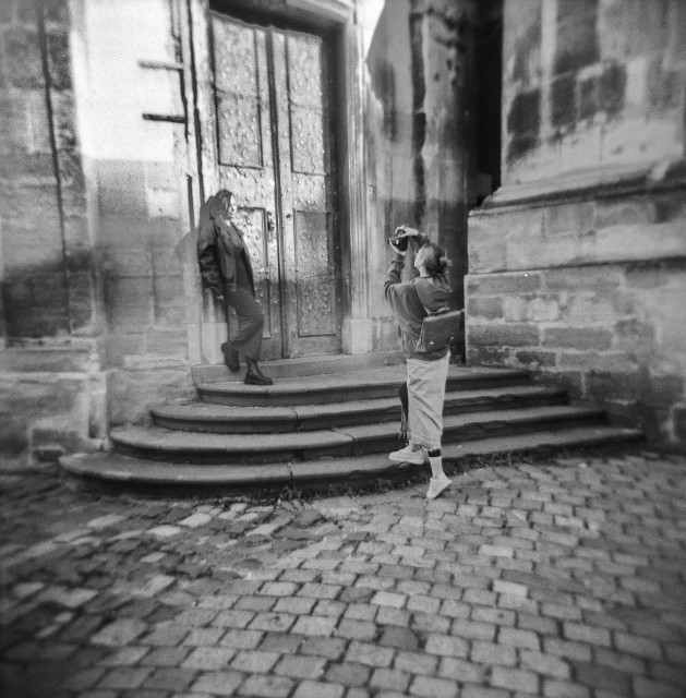 The image is a black-and-white photograph depicting a scene with two young women near an old, textured doorway. The setting appears historical or vintage, as suggested by the cobblestone pavement and ornate architectural details of the building. 

One girl, standing on the stone steps near the door, is leaning casually against the wall,  posing for a photograph. She is dressed in dark clothing, exuding a relaxed, moody vibe. The other girl, positioned slightly lower on the steps, is holding a camera and taking a photo of the first person. This photographer is wearing a light-colored outfit with a city backpack on her back. 

The photograph has a soft, dreamlike quality, enhancing its nostalgic atmosphere.