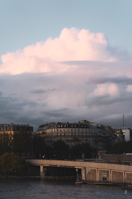 The image depicts an urban skyline at dusk, with a dramatic cloud formation overhead. A bridge spans the body of water in the foreground, and buildings with a mix of architectural styles can be seen in the background. The warm lighting and atmospheric clouds create a moody, picturesque scene.