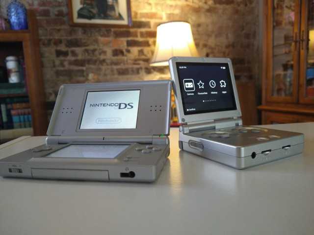 A silver DS Lite and silver RG35XXSP seated side by side on top of a white surface with a brick wall and bright lamp in the background