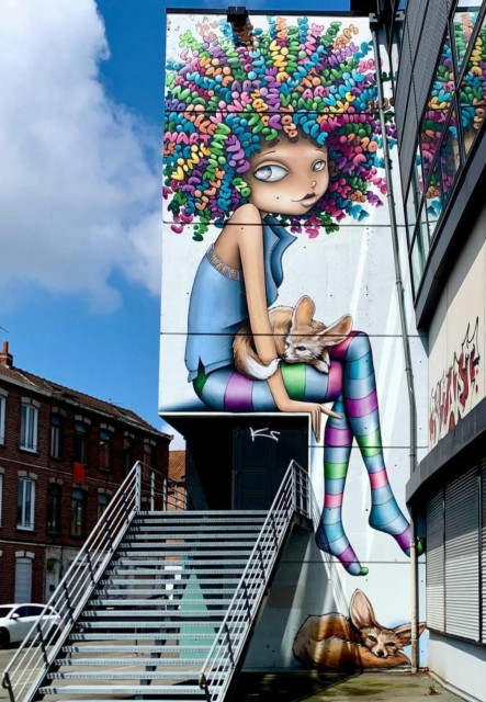 Streetartwall. An enchanting mural of a girl in comic style is sprayed on a narrow exterior wall of a modern building above a staircase. The girl is wearing a blue top and long stockings in bright rainbow colors. She has big eyes, a small nose and a small mouth. On closer inspection, her curly, wispy hair is made up of beautifully written colorful words, e.g. the names of the artists Vinie, Akhine and the organizer Collectif Renart, 2020, etc. The girl is sitting with her legs crossed over the entrance to the house and the stairs, holding a small fox on her lap. Another fox is lying on the floor next to the stairs. A beautiful splash of color that brightens up the whole area. 
Info: The girl figure is from "Vinie", the foxes from "Akhine".