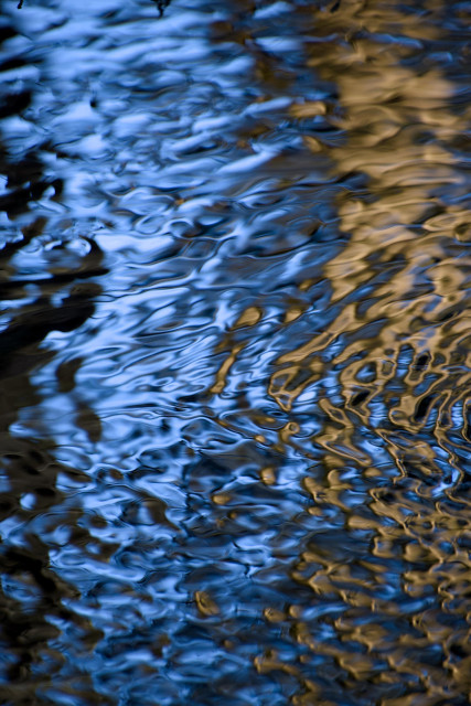 Colour photograph taken with a long lens and fast shutter speed of the reflections of bankside trees and the bright blue sky in a shallow stream, where the ripples have created beautiful patterns