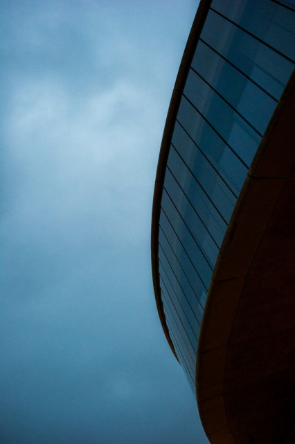 Photo loooking up at a blue-white cloudy sky. On the right, a curve of thin windows separated only by black lines swoop from the top right to the bottom left, standing out from a building off-camera.