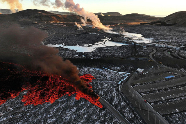 Lava is flowing to the right where there is a big empty parking lot except for a few cars.   A protective barrier is also seen and it looks like to machines are working on it. 