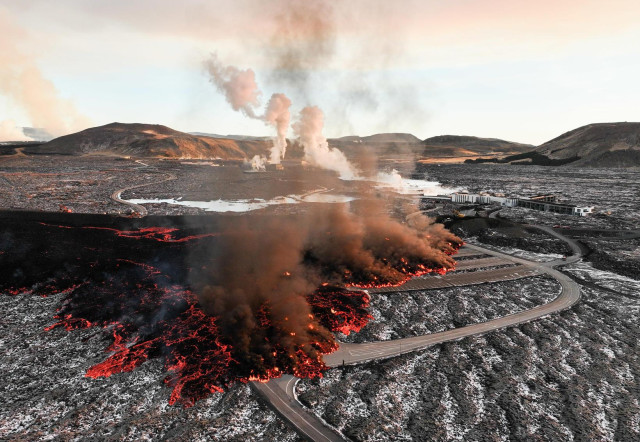 View of the lava flow from above, lava is flowing over a big parking lot and on the road nearby. In the background is a big barrier protecting a geothermal powerplant.