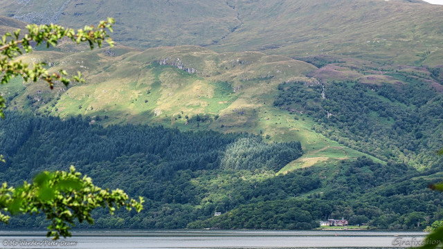 A photo of a heavily wooded mountainside, the upper slopes are green with grasses and more scattered trees. Dappled sunlight from the left is playing across the contours, but the sky is out of shot. At centre right, a waterfall can be seen between the trees. Just above it and nearer to the centre, bare rock is visible. Down at the still waterline is a cluster of buildings and another, smaller one, to its right and nearer the centre. In the foreground is an out-of-focus tree branch covered in green leaves.