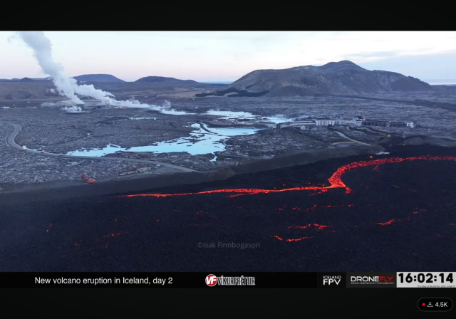 Screenshot from livestream. A huge lava flow are running alongside a barrier. Behind the barrier are blue water and buildings. To the left a red excavator is working on the barrier. 