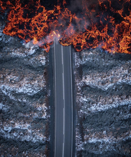 A road seen from above, on the sides of the road is old lava with a thin cover of snow, fresh lava is moving in from the top slowly swallowing the road.