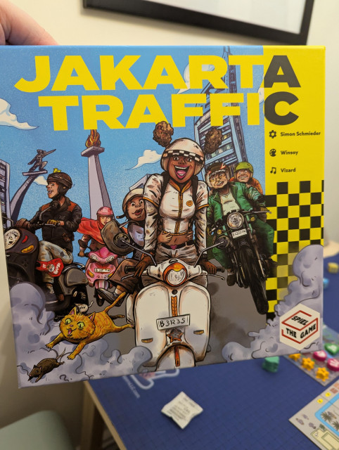 A brightly-colored boardgame box for Jakarta Traffic, depicting several enthusiastic scooter delivery people and an orange cat chasing a rat