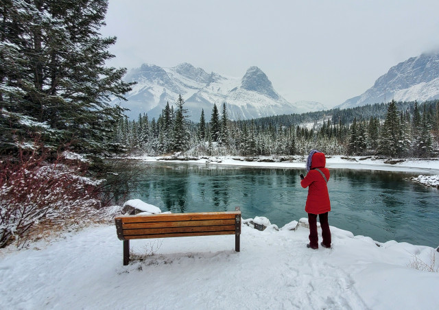 A cold snowy late November day alongside the Bow River at Canmore Alberta in the Canadian Rocky Mountains, just outside of Banff National Park. A brown wood bench faces the river and mountains beyond, which are slightly obscured by the overcast sky and falling snow. A woman in a red coat stands beside the bench observing the view. 