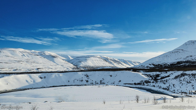 A photo of a snow scene with a river curving through the foreground. A range of mountains fills the horizon and disappears into a valley behind another foreground peak at centre right. The sky above has scattered cloud but is deep cold blue and cyan. The sunlight is from the right. The snow lying on every surface is completely untrammelled.