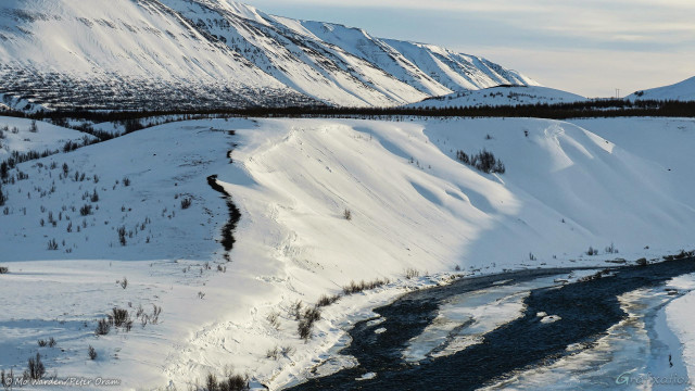 A photo of a snow scene, and the same river except this time in close-up. The untouched snow on the riverbanks is shining and silky, long shadows are being thrown from the right by strong sunlight. A line of mountains on the horizon has a cloud-bank above it. The river water is dark and has large areas completely frozen, most of which are also covered in snow.