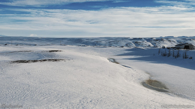 A photo of a snow scene, under a cloudy but blue sky. The horizon is hilly and a farm building in the centre right lends scale. The foreground is a smooth snow-field, its surface slightly pitted and with dark areas of rock showing through. On the right, in a depression, part of the river is visible as a few slightly thawed patches next to a frozen and snowed-over surface. The end is a river bank so this would be a pool when thawed.