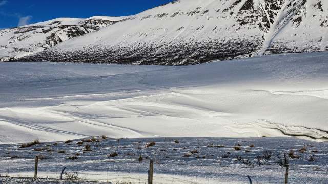 A photo of a snow scene but, this time, from further away from the river so that its surface can't be seen. The horizon is obscured by large mountainsides and only a small window of blue sky is visible top left. The foreground is a fenced field. Between this and the mountains are beautifully sculpted silken snowdrifts, lit from almost directly behind the viewpoint. The snow is clean and untouched, shadows on its surface are the only sign that it's folded into gorgeous convoluted pleats.