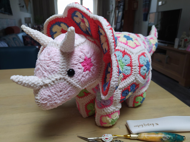 patchwork triceratops dinosaur made from crochet african flowers, the head is off-white with light pink centers and darker pink blushes and off-white horns, the body and frill are off-white with colorful centers and the tail has a jaunty upward curve